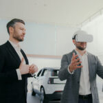 Two Caucasian men in modern showroom. Male wearing VR headset and gesturing with his hand. Virtual environment. Consultant wearing formal suit observing and giving instructions or explanations.